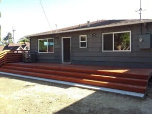 A single-story gray house with a wooden deck features two large windows and a central door. The yard, primarily bare soil, offers potential for creative landscaping to complement the inviting decks.