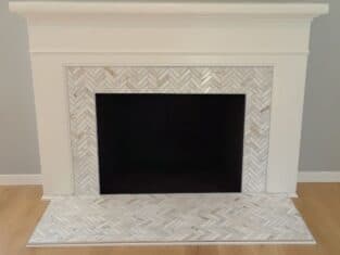 White fireplace with a herringbone-patterned tile surround against a gray wall and light wood floor, seamlessly complementing the elegant bathrooms nearby. Two wall outlets are positioned above the mantle.