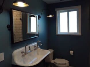 This bathroom, a prime example of residential construction, features blue walls, a double sink, large mirror, toilet, and window. Wall-mounted lights illuminate the space perfectly.