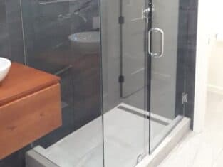 A sleek glass shower enclosure with a metal handle and hinges stands against dark tiled walls, enhancing the modern bathroom. A wooden vanity with a white basin is partially visible to the left, creating a harmonious blend of natural textures.