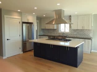 Modern kitchen with stainless steel appliances, a large island with gas stove, white cabinets, black base, range hood, and subway tile backsplash. Light wood flooring complements the craftsmanship found in every corner of this residential construction masterpiece.