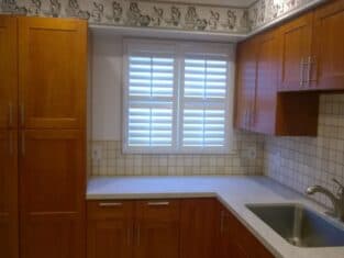 The kitchen features wooden cabinets, white countertops, a tiled backsplash, and a stainless steel sink. Above the counter, a window with shutters adds charm, while a decorative border graces the wall near the ceiling—a nod to sleek commercial construction aesthetics.