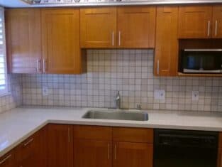 Kitchen with wooden cabinets, a stainless steel sink, microwave, and dishwasher. Countertops are light-colored, the backsplash tiled in a grid pattern. A window with shutters complements the room’s aesthetic. Perfect for homeowners considering a full remodeling project to match their style.