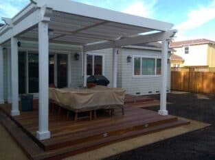 A backyard patio features a wooden deck with a white pergola, outdoor furniture covered with a tarp, and a barbecue grill, highlighting the charm of recent residential construction. A fenced yard and neighboring houses are visible.