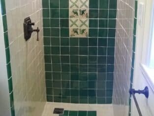 Shower with green and white tiles, featuring a floral pattern strip in the center and a matching floor accent—perfect for a remodeling project. Black showerhead and handle on the left wall add a sleek touch.