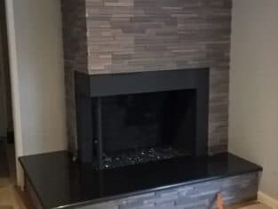 Stone fireplace with black mantel and hearth is the focal point of this partially unfinished residential construction, complete with bare floors, gray walls, and a recessed ceiling light.