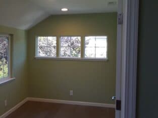 A small, empty room with green walls awaits its transformation, ideal for remodeling. It features three small windows on one side and a larger window on another, complemented by a wooden floor.