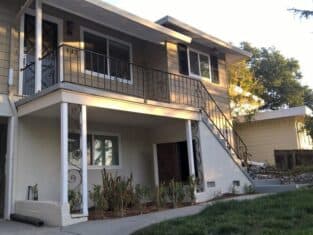 A two-story beige house showcases the charm of residential construction, featuring a staircase leading to an upper balcony. On the ground level, a garage and small garden are visible, surrounded by lush bushes and trees in the yard.