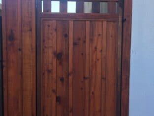 A wooden gate with vertical slats and a small open section at the top stands elegantly between two walls, seamlessly complementing nearby fences.