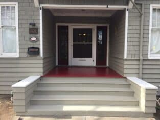 Front porch of a house with gray siding, red floor, and white-trimmed windows and door, showcasing expert residential construction. A house number is displayed on the left side near a mailbox.