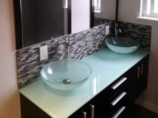 This elegantly designed bathroom features two glass vessel sinks on a light green countertop, complemented by dark wood cabinets and a mosaic backsplash. Perfect for modern residential construction, it includes two mirrors with three-bulb lights above, nestled under small windows.