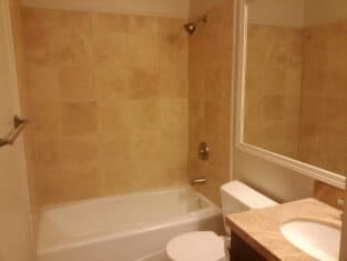 A serene bathroom featuring beige tiled walls, a bathtub and shower, a white toilet, and a sink with a matching beige countertop. The space is completed by a large mirror above the sink. Ideal for those who cherish elegant bathrooms with timeless design.