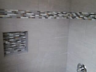 Shower with gray tiles and mosaic accent strip features a built-in niche. Contains a showerhead, faucet, and bathtub. Perfect for those considering bathroom remodeling projects to add style and functionality.