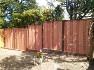 Wooden fence with vertical panels graces the garden, bordered by lush trees and shrubs. A pot with vibrant orange flowers sits in front, perfectly complementing the serene outdoor remodeling effort.