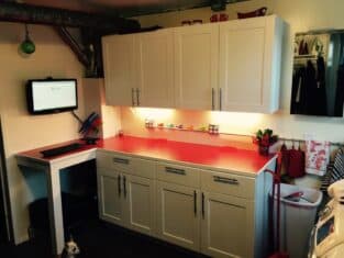A small kitchen featuring white cabinets and vibrant red countertops hosts a wall-mounted computer and an array of colorful utensils and cups. A curious dog peeks from the bottom, adding charm to this cozy space, reminiscent of those unexpected joy found beyond decks or fences.
