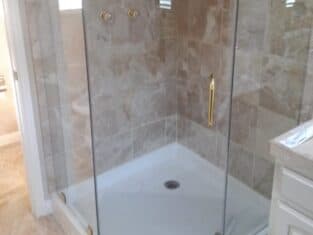 This elegant residential bathroom showcases a glass shower enclosure with gold trim and marble tile walls. It features dual shower controls, a corner shelf with a red bottle, and a pristine white shower base for a luxurious bathing experience.