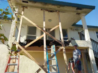 A person engaged in commercial construction stands near a partially collapsed balcony supported by wooden beams and ladders, wearing a cap, safety goggles, gloves, and holding a tool in front of a two-story building.