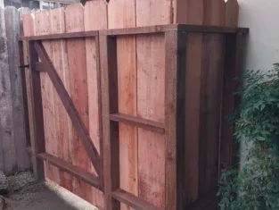 Wooden gate with vertical planks and a diagonal support beam, seamlessly blending into a residential construction setting. Set within a fence structure beside the house, it is complemented by green foliage visible on the right side.