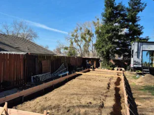 A rectangular wooden frame for a backyard foundation hints at remodeling ambitions, surrounded by a tree, fence, and RV under a clear blue sky.