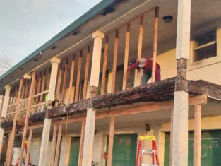 Two people work on the balcony of a two-story building. One stands on the balcony, repairing wooden beams, while the other stands near the house exterior. Red ladders are visible as they focus on both deck and fence restoration.