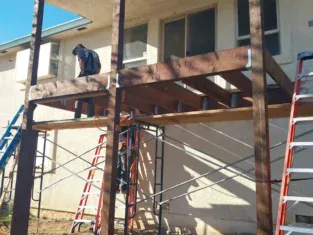 Two people work on constructing a wooden deck attached to a house, with scaffolding and ladders providing access and support. The project enhances the outdoor space, much like adding new bathrooms inside enriches the home’s interior functionality and comfort.