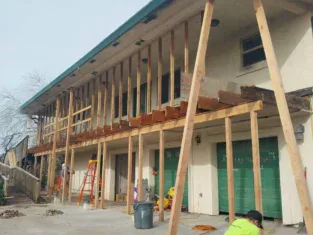 House renovation in progress with wooden beams supporting the upper deck. A worker in a yellow jacket is seated on the ground, surrounded by tools and equipment, reflecting the precision and skill usually seen in commercial construction projects.