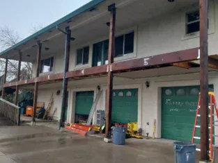 The commercial construction site features a building facade under renovation, with exposed wooden beams and materials scattered on the wet pavement. Green garage doors lie below two levels of windows, set amidst ladders and tools, indicating an extensive remodeling process in progress.