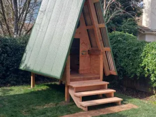 A small A-frame wooden playhouse with a green metal roof stands on a lawn, surrounded by trees and bushes. It hints at the charm of residential construction, with its short staircase leading up to the entrance.