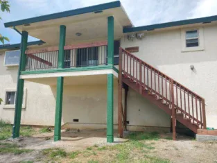 Exterior view of a beige building with green pillars, showcasing residential construction, features a wooden staircase leading to an elevated entrance. Lush grass surrounds the base, adding charm and character.