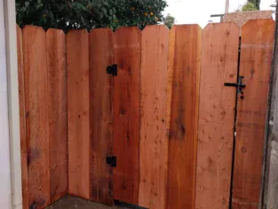 A wooden fence gate with metal hinges and a latch, nestled among trees and a white wall, hints at residential construction. A black drainage pipe is visible on the ground.