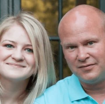 A woman with long blonde hair and a man with a shaved head pose together in front of a window, showcasing the stunning results of their recent remodeling project.